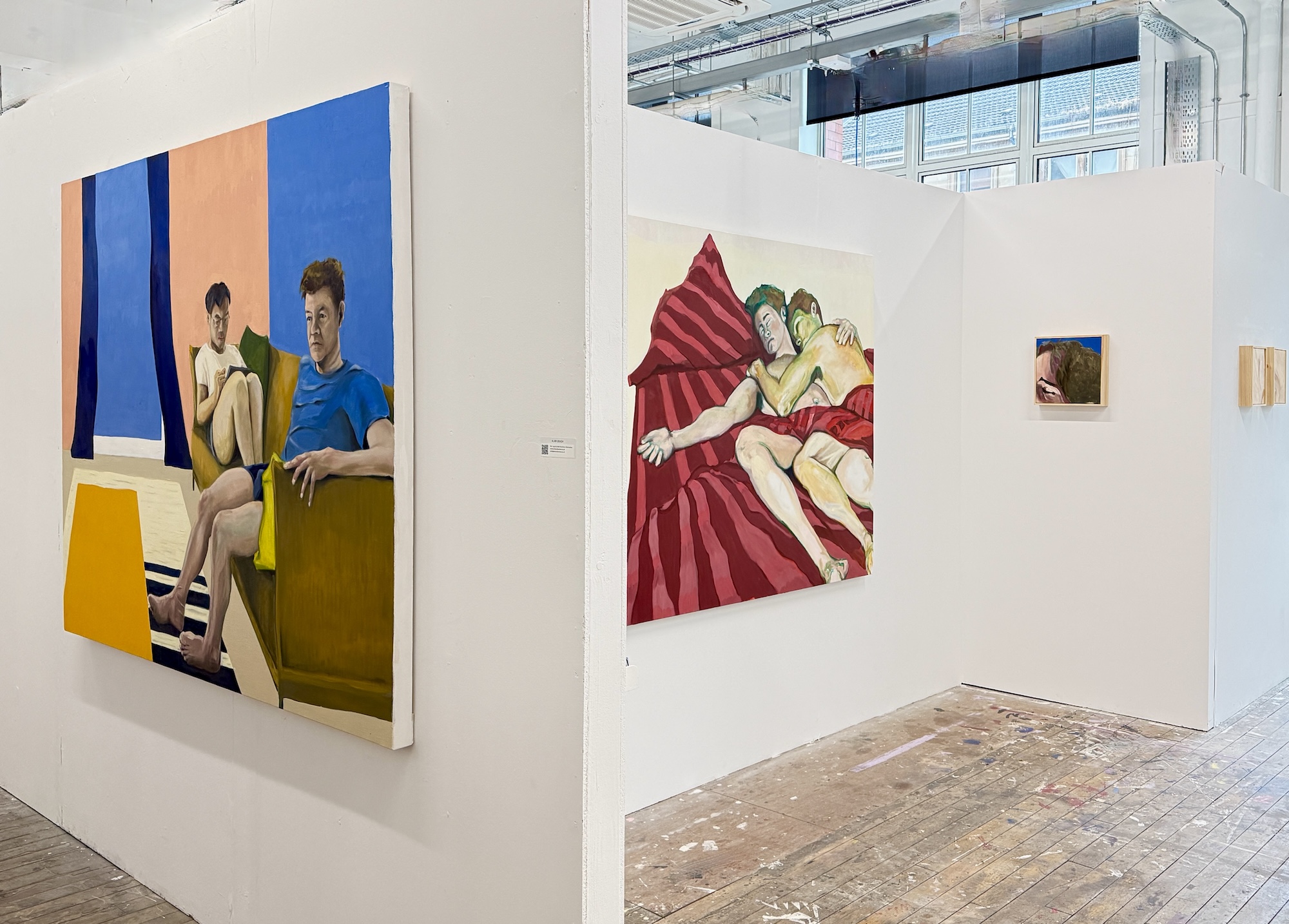 Installation view: a seated couple on a sofa, with a large red bed painting in the neighbouring space.